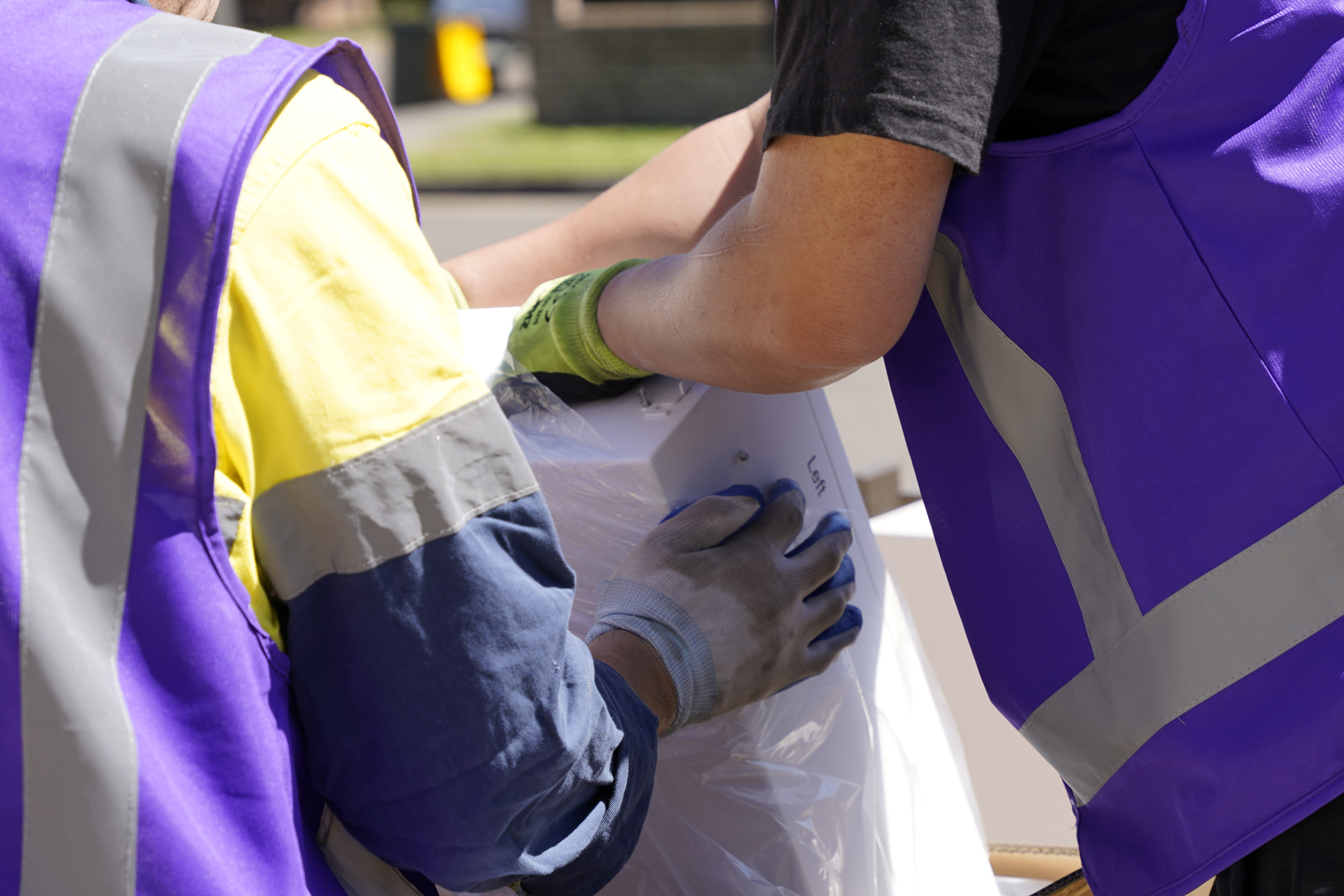 Two workers in purple vests are lifting a large package together, focusing on their coordinated effort.