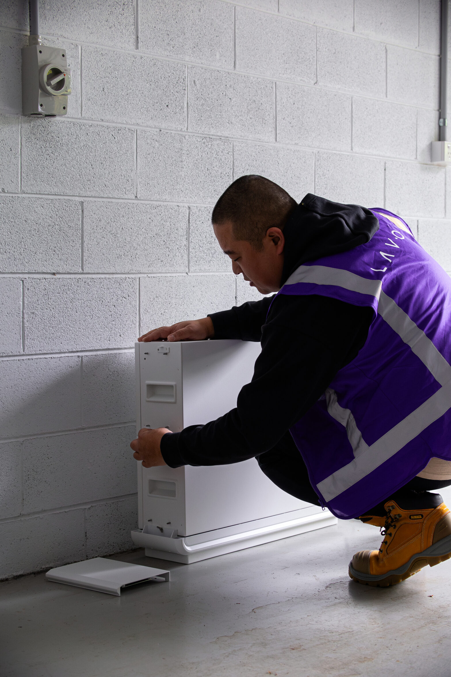 A worker in a purple vest installs a heating unit against a gray wall in a well-lit space.
