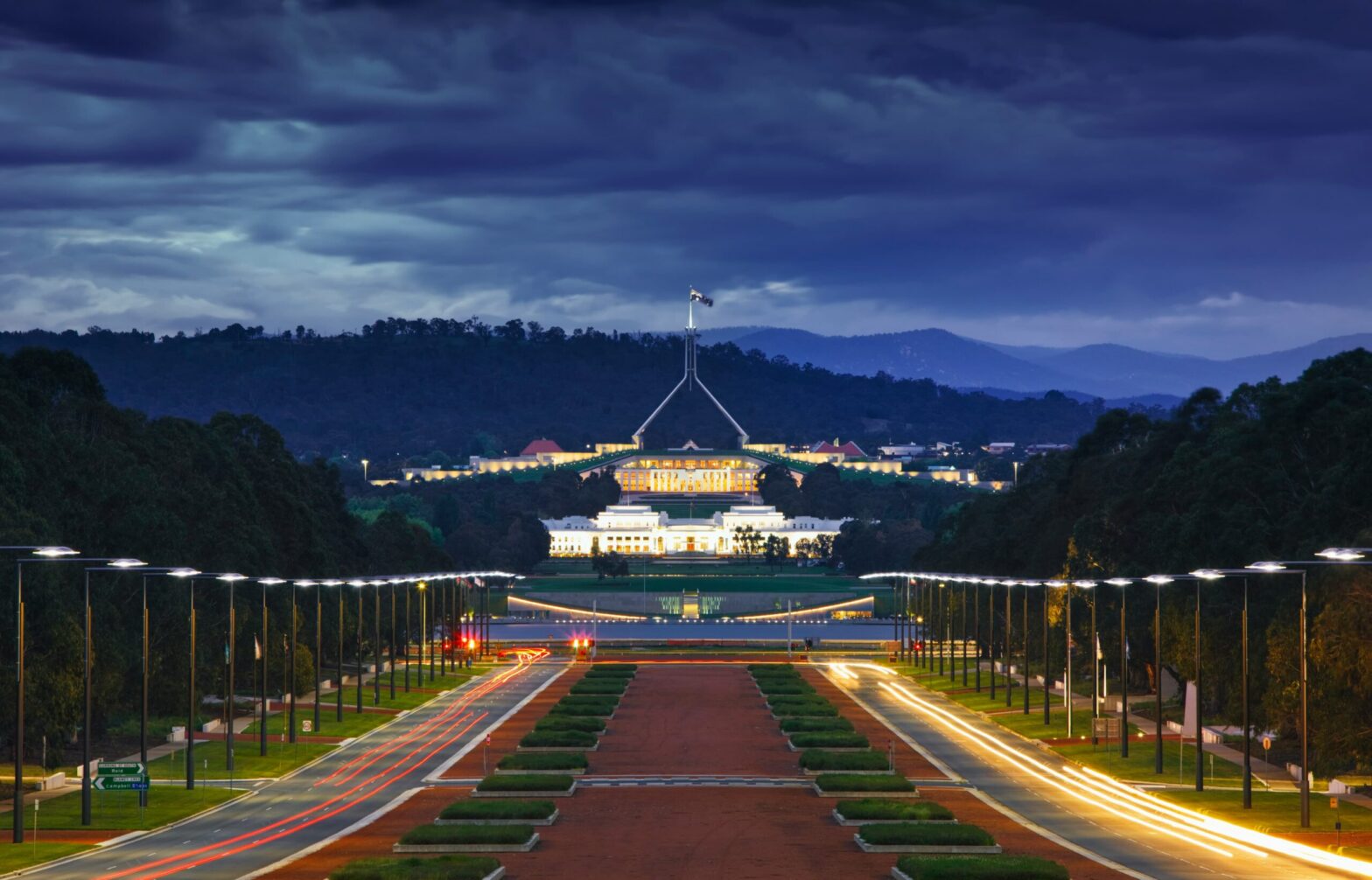 Australian Parliament House in Canberra illuminated at night with the road leading up to the building under a dramatic cloudy sky
