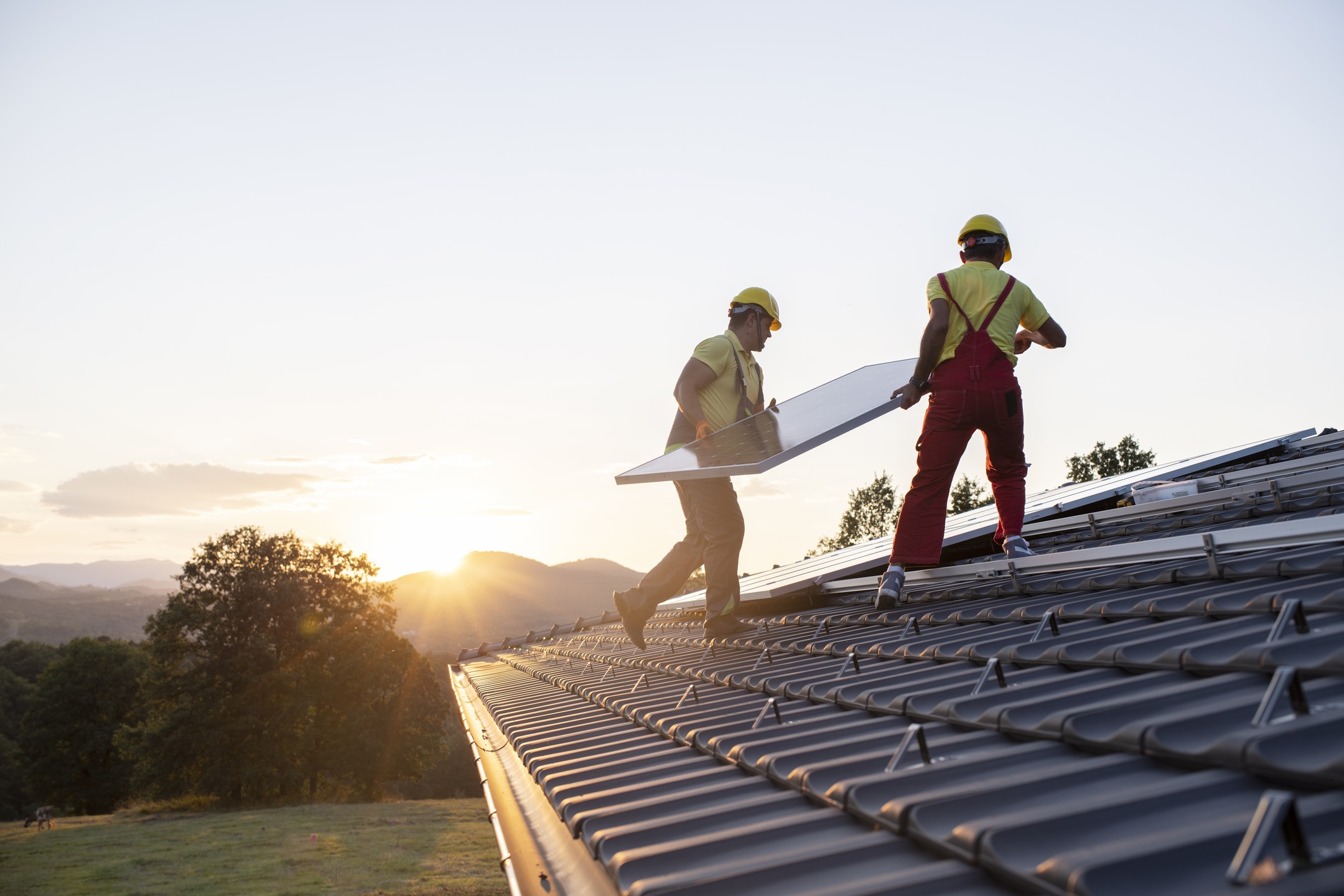 Two workers in safety gear installing solar panels on a rooftop at sunset.