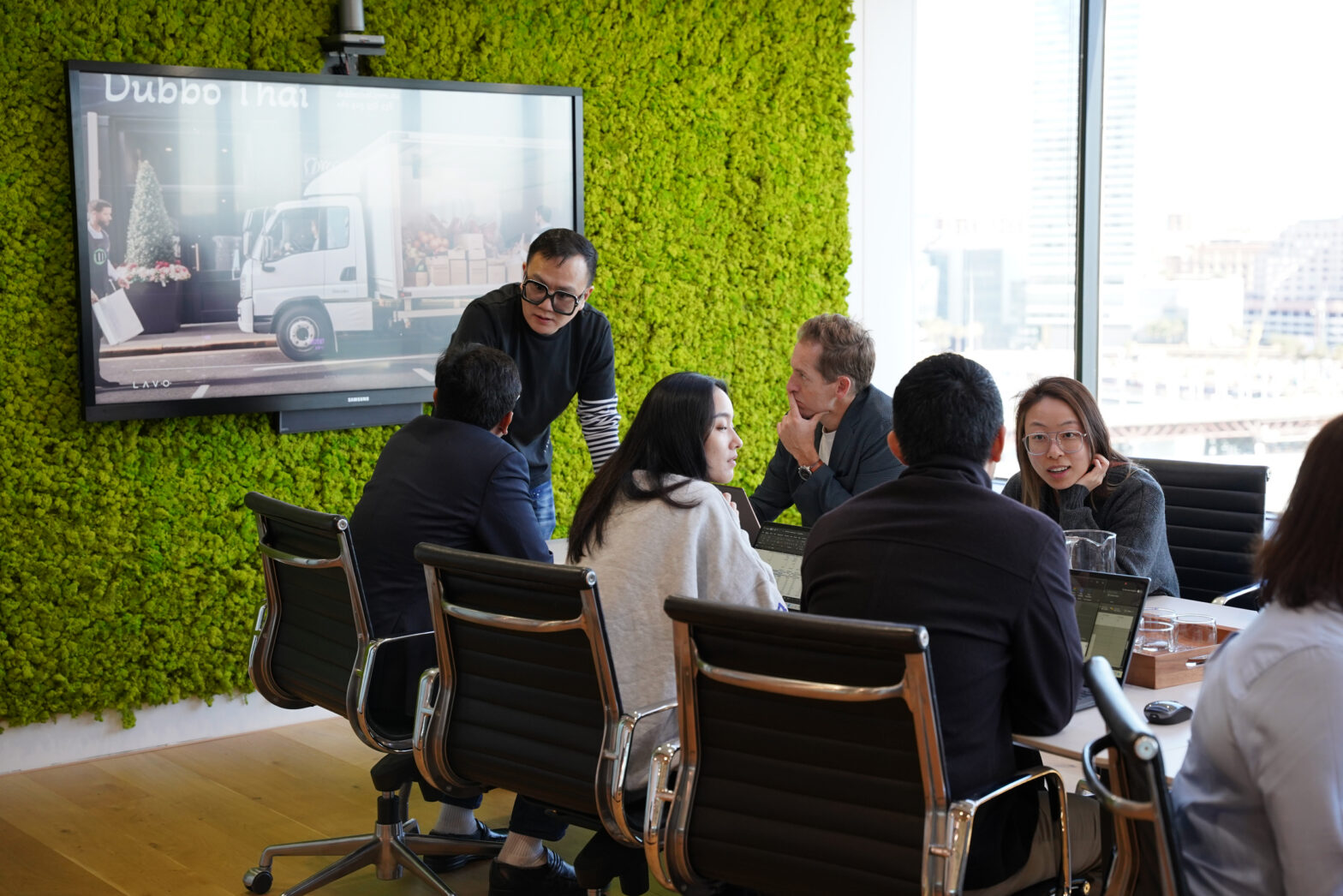 A group of professionals collaborates in a modern meeting room with a green wall and a screen displaying a truck image.