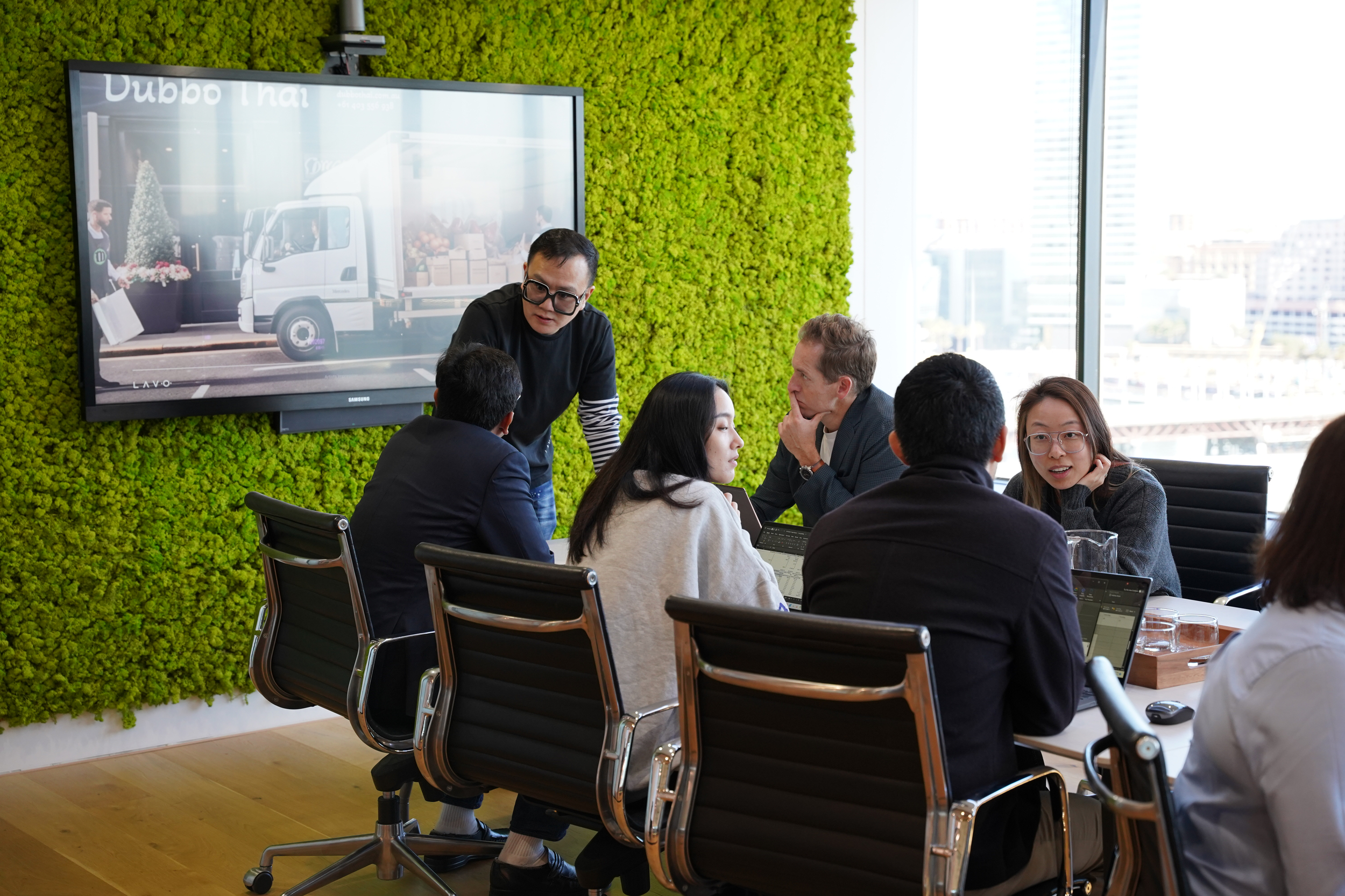 A group of professionals collaborates in a modern meeting room with a green wall and a screen displaying a truck image.