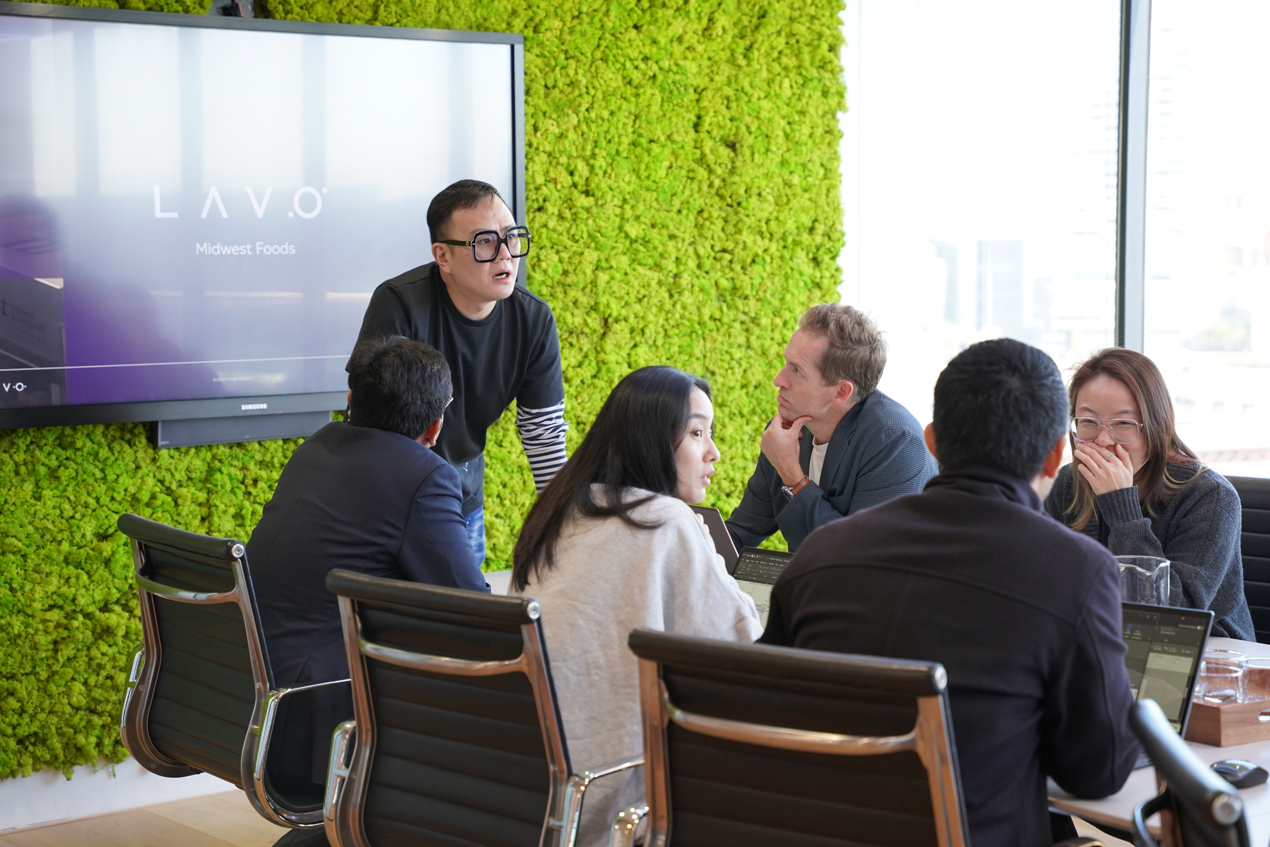 A group of professionals meets around a table, with a presenter in front of a green wall and a screen displaying "LAVO".