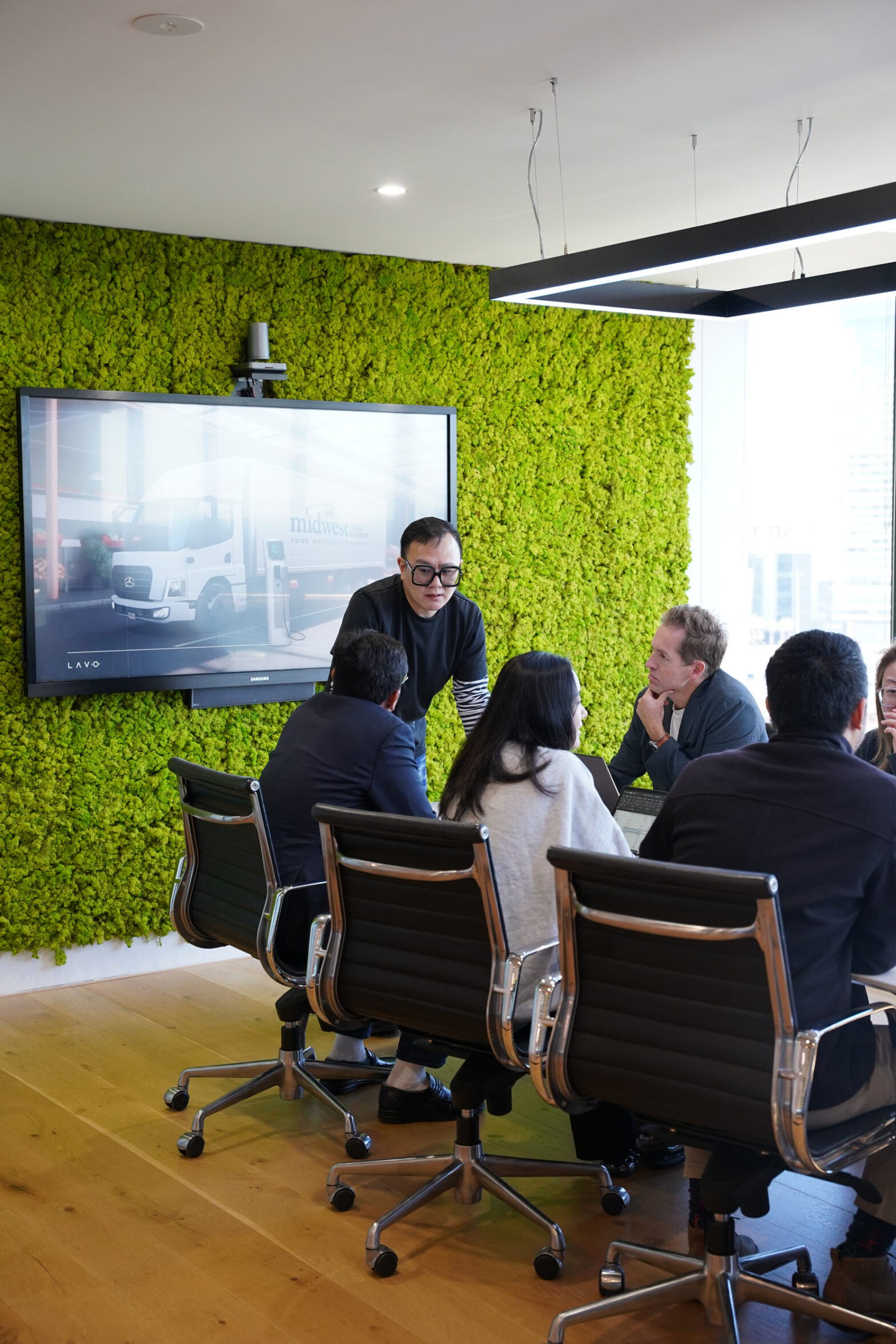 A group of professionals sits around a table in a modern meeting room with a green wall, discussing a presentation on a screen.
