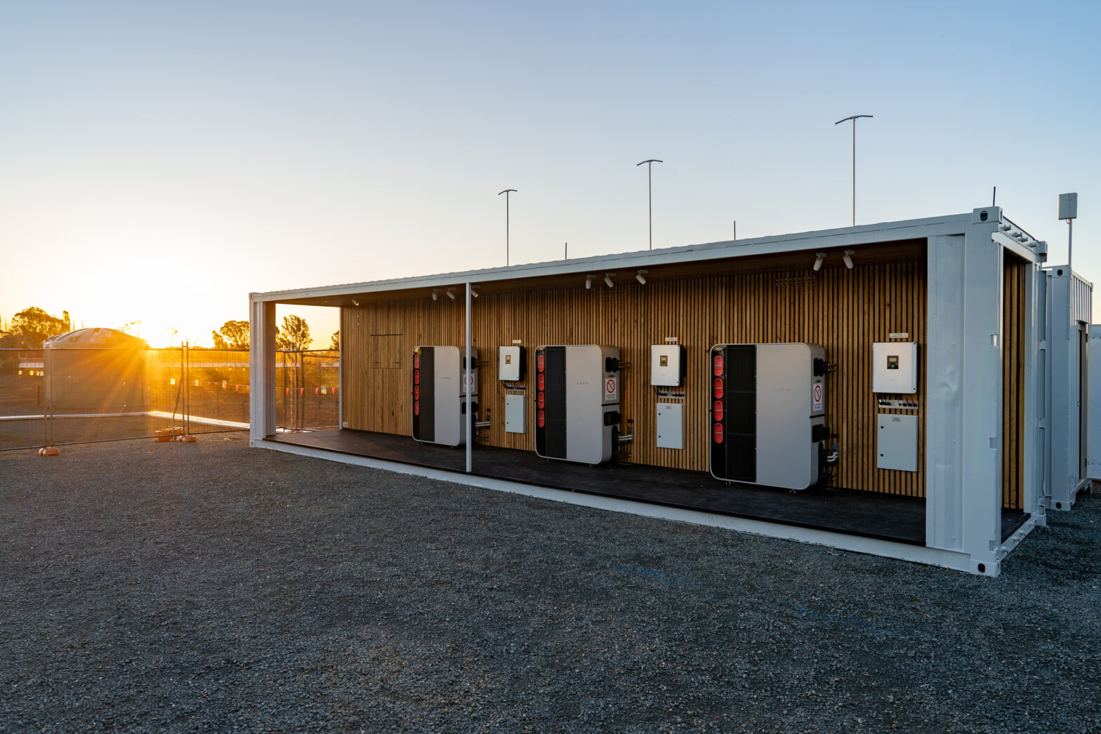 A modern wooden structure with several vending machines illuminated by the sunset in a gravel area.