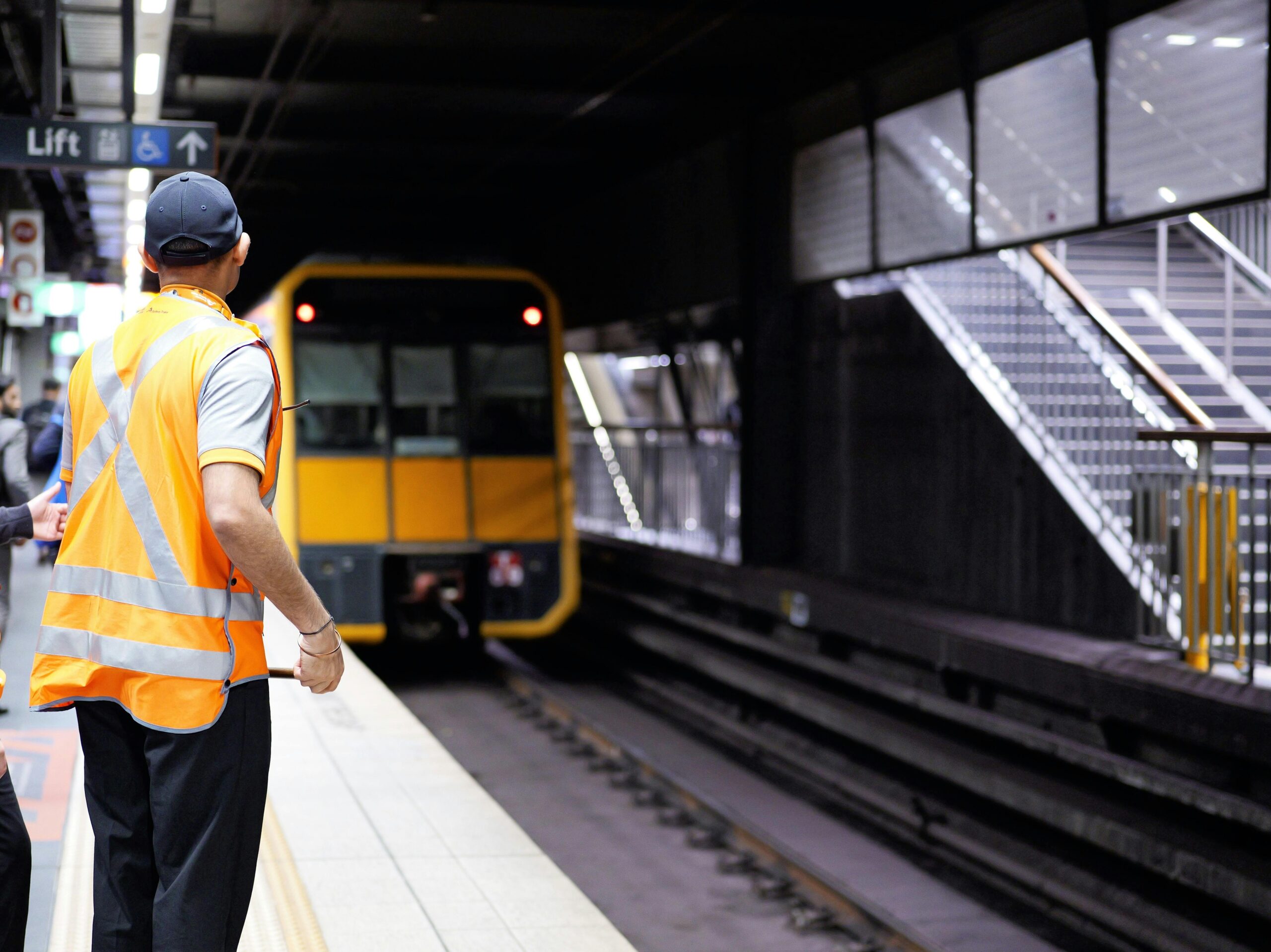 Railway worker in a high-visibility vest standing on a platform as a yellow train approaches